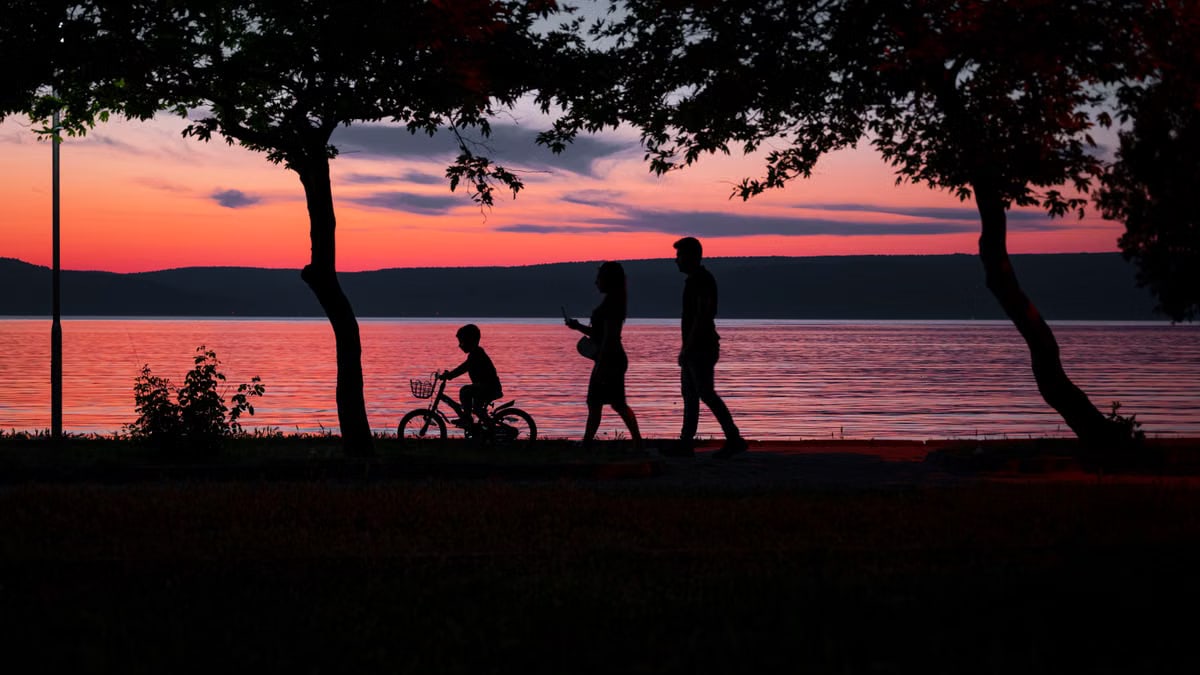Silhouette of a family walking and biking by a lake at sunset, framed by trees.